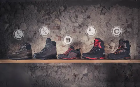 Fünf Wanderschuhe verschiedener Kategorien, darunter Kategorie B, stehen auf einem Regal vor einer Steinwand.