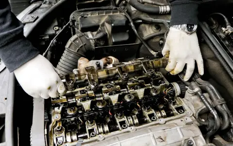 Mechanik w białych rękawiczkach sprawdza wałek rozrządu, szukając objawów zużycia. Widoczne są elementy silnika i przewody.