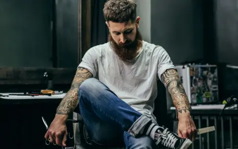 Mężczyzna z brodą i tatuażem na biceps siedzi w fotelu, trzymając nożyczki.
