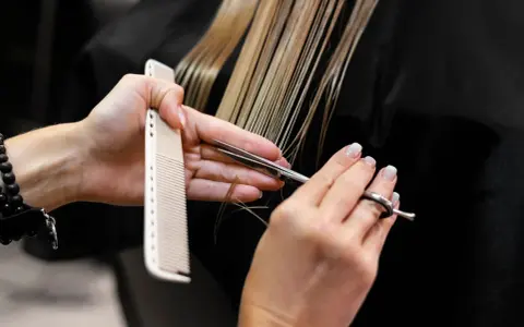 Manos de estilista cortando cabello rubio con tijeras y peine. Decisión: cortar el pelo en seco o mojado.