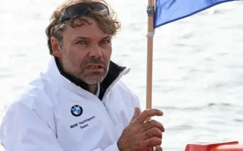 Markus Wieser, mit zerzaustem Haar und Sonnenbrille auf dem Kopf, trägt eine weiße BMW Yachtsport Team Jacke. Er blickt konzentriert auf das Wasser.