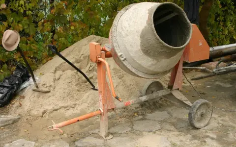 Betoniarka 200l czeka na piasek i cement. Ile cementu na betoniarkę 200l? Obok łopata i kapelusz.