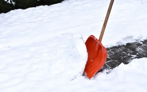 Pomarańczowa łopata do śniegu wbita w zaspę na ośnieżonym chodniku.