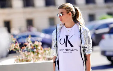 Kobieta w srebrnej kurtce CK damskiej i białej koszulce z logo Calvin Klein Jeans.