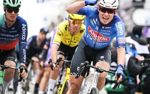 Radfahrer im blauen Trikot von Alpecin-Deceuninck jubelt nach einem Sieg, während andere Fahrer im Peloton folgen.