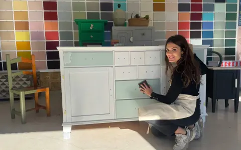 Mujer sonríe mientras trabaja en un mueble, aplicando pintura blanca. Aprende como pintar un mueble oscuro en blanco sin lijar.