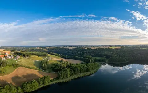 Malownicze **lasy w Polsce** otaczają spokojne jezioro. W oddali widać wieś z czerwonymi dachami, pola i pagórki.