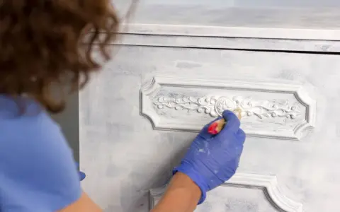 Mano enguantada aplica pintura a muebles pintados a la tiza en dos colores, dando un acabado desgastado y elegante.