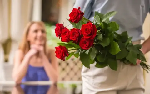 Mężczyzna z bukietem czerwonych róż za plecami, zbliża się do uśmiechniętej kobiety. To piękny gest, jak na 60 lat po ślubie, jakie to gody!