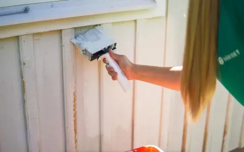 En person med långt blont hår målar en vit träfasad med en bred pensel. Det är dags att måla av huset för sommaren.