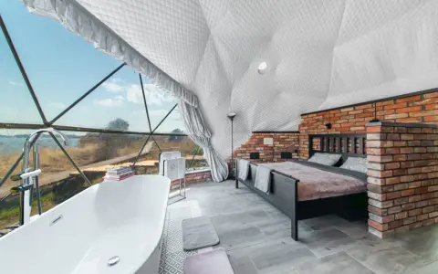 Luksusowy glamping nad jeziorem: wanna z widokiem, łóżko i ceglana ściana w kopułowym wnętrzu.