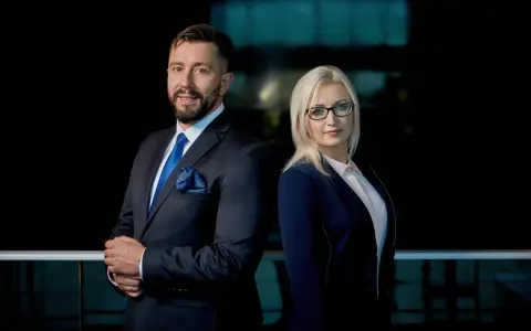 Para prawników w eleganckich garniturach, mężczyzna i kobieta, stojący na tle ciemnego biura.