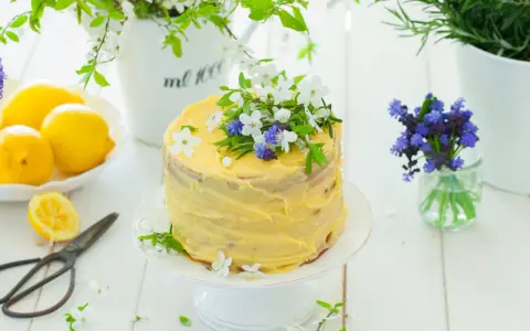 Słoneczny tort cytrynowy z kremem lemon curd, ozdobiony świeżymi kwiatami i ziołami. Obok leżą cytryny i stare nożyczki.