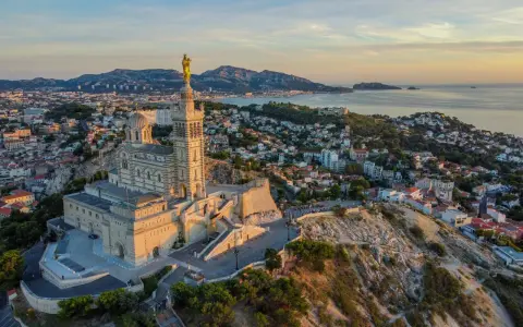 La basilique Notre-Dame de la Garde veille sur Marseille, baignée par la lumière dorée du soleil couchant. La mer et les collines environnantes s'étendent à perte de vue.