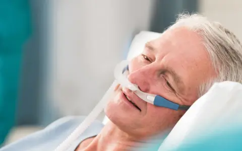 Älterer Mann mit Nasenkanüle, der entspannt im Bett liegt. Die Sauerstoffzufuhr mit **high flow** hilft ihm beim Atmen.
