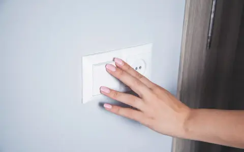 Main d'une femme aux ongles roses actionnant un interrupteur blanc, prêt pour le branchement d'une lampe de chevet.