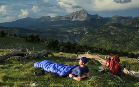 Mann liegt in blauer Biwakschachtel im Freien, umgeben von Berglandschaft. Rucksack und Wanderstöcke daneben.