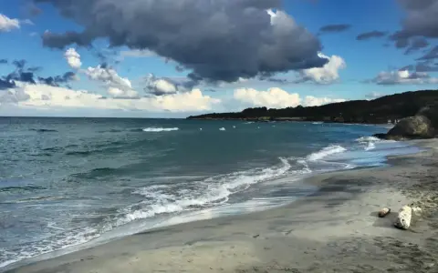 Onda che lambisce la spiaggia sabbiosa dell'Isola d'Elba, con il mare azzurro e nuvole scure nel cielo.