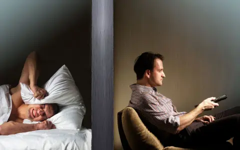 Hombre en cama tapa oídos con almohada, sufriendo por vecinos ruidosos. Al otro lado, otro hombre ve TV.