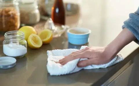 Mano limpiando una encimera con un paño blanco, junto a un frasco de bicarbonato y limones.