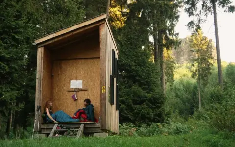 Zwei Personen entspannen sich in einer einfachen Holzhütte, bereit für wild camping. Umgeben von hohen Bäumen und üppigem Grün.