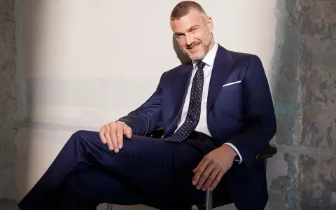 Uśmiechnięty mężczyzna w granatowym garniturze, z krawatem o klasycznej szerokości, siedzi w fotelu.