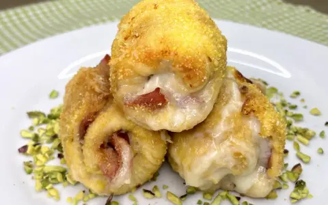 Tre involtini di pollo dorati e ripieni di formaggio filante e prosciutto, adagiati su un letto di pistacchi tritati.
