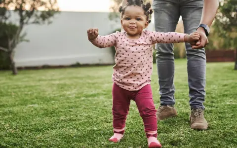 Ein kleines Mädchen mit lustigen Zöpfen macht seine ersten Schritte im Gras, gehalten von einer erwachsenen Hand. Wann laufen Babys? Heute ist ein guter Tag dafür!