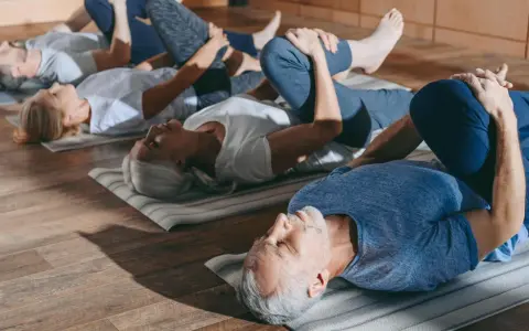 Grupa seniorów 60 plus ćwiczy na macie. Wszyscy leżą na plecach, przyciągając jedno kolano do klatki piersiowej.