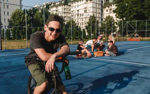 Uśmiechnięty chłopak na rowerze z butelką piwa, obok grupa znajomych. Może to być scena, która zakończy się mandatem za jazdę rowerem po alkoholu.