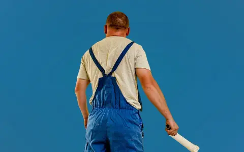 Mężczyzna w niebieskich ogrodniczkach i jasnej koszulce trzyma wałek malarski.