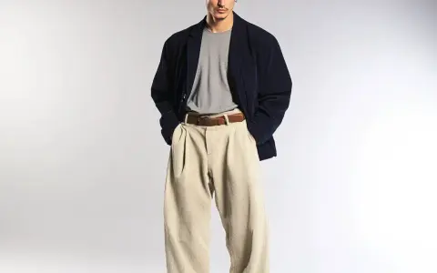 Uomo con spezzato giacca blu scuro, maglietta grigia e pantaloni beige a vita alta.