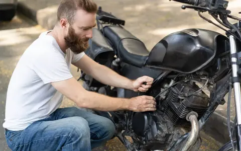 Uomo con barba lavora sul motore di una moto. Controlla i cavi, forse per il tempo di ricarica batteria moto 12v.