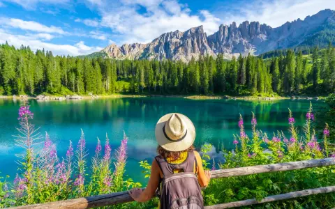 En kvinna med hatt och ryggsäck njuter av den vackra **Dolomiterna sommar**-utsikten över en turkos sjö, omgiven av skog och berg.