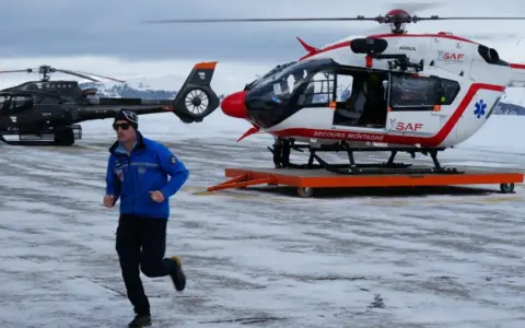 Ratownik w niebieskiej kurtce biegnie na tle dwóch helikopterów w Austrii, gotowych do akcji po lawinie.