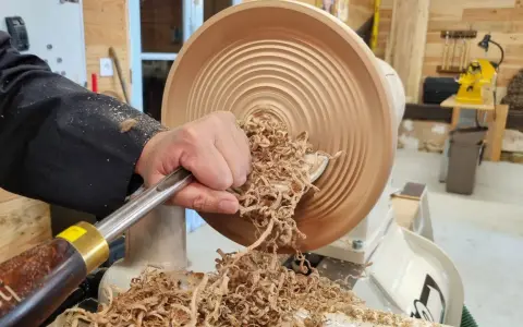 Main d'un artisan façonnant un objet en bois sur un tour. Les copeaux volent, témoignant du travail de tournage sur bois.