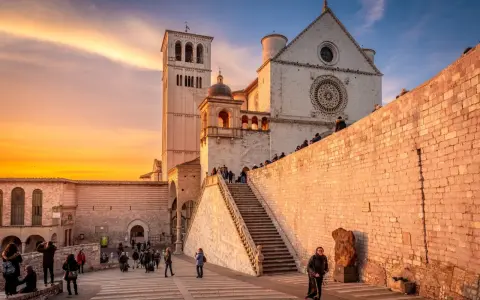 Assisi cosa vedere: la Basilica di San Francesco al tramonto, con la sua imponente facciata e la torre campanaria, illuminata da una calda luce dorata.