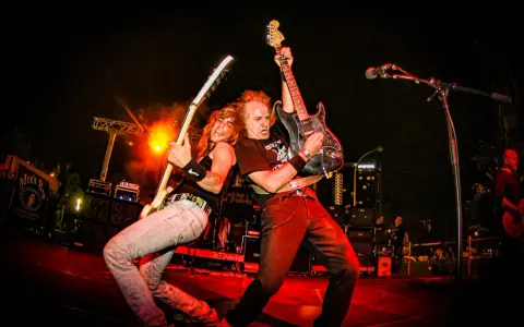 Dos guitarristas de las ruedas grupo musical tocan con pasión en un escenario iluminado por luces rojas y naranjas.
