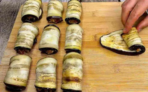 Nove involtini di melanzane pronti, uno viene arrotolato su una fetta di melanzana grigliata.