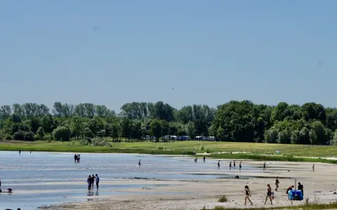 Menschen genießen einen sonnigen Tag am Strandbad Eldena. Einige baden im flachen Wasser, andere spazieren am Ufer entlang.