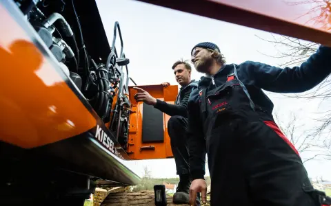 Dwóch mechaników w kombinezonach przeprowadza konserwację maszyn budowlanych. Jeden z nich wskazuje na silnik.