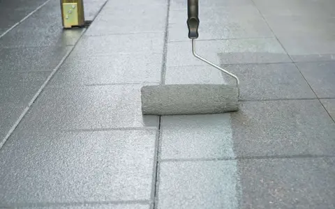 Ein Farbroller streicht graue Pflastersteine. Die rechte Seite ist frisch gestrichen, die linke Seite noch ungestrichen.