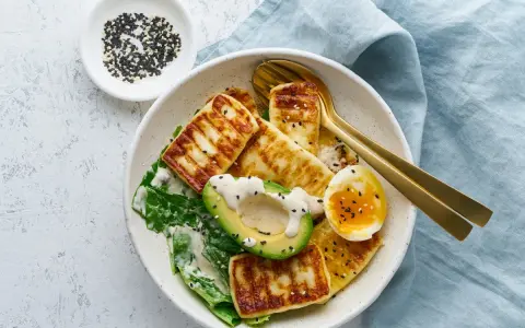 Szybki keto obiad: grillowany ser halloumi, awokado, jajko na miękko i zielone liście, posypane sezamem.