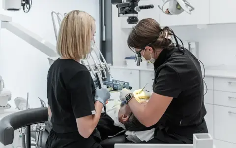 Dwie dentystki pracują przy pacjencie. Jedna używa mikroskopu, druga trzyma narzędzie. To obrazek pokazujący, jak zostać dentystą.