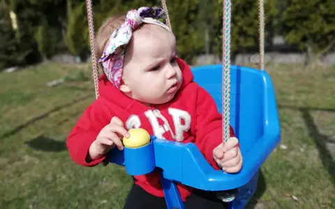 14 miesięczne dziecko w czerwonej bluzie z kapturem i kolorowej opasce na głowie siedzi na niebieskiej huśtawce na zewnątrz.