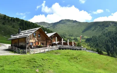 Malga Alto Adige, un rifugio in legno con tavoli all'aperto, circondato da verdi pendii e montagne boscose sotto un cielo azzurro con nuvole bianche.