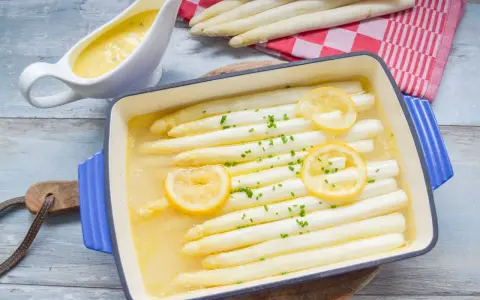 Weißer Spargel in einer Auflaufform mit Zitrone und Kräutern, daneben eine Sauciere mit Sauce.