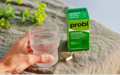 En hand håller ett glas vatten bredvid en ask med Probi Original. Förpackningen förklarar vad innehåller probiotika: levande mjölksyrabakterier.