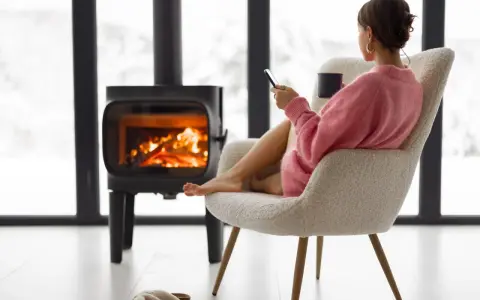 Femme assise près d'un poêle à bois, tenant une tasse et un téléphone.