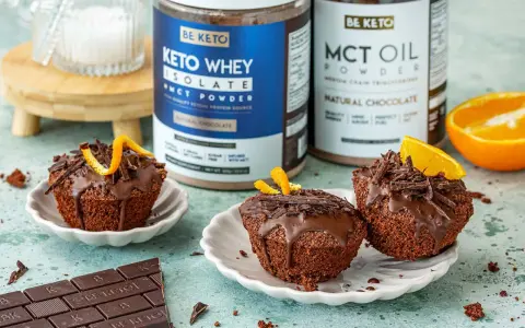 Pyszne keto muffiny z mąki migdałowej, polane czekoladą i ozdobione skórką pomarańczową. Obok stoją puszki z odżywką białkową i olejem MCT.